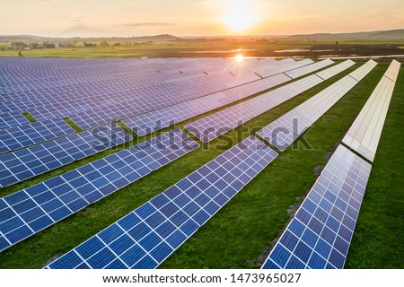 Blue solar photo voltaic panels system producing renewable clean energy on rural landscape and setting sun background. Royalty-Free Stock Photo #1473965027