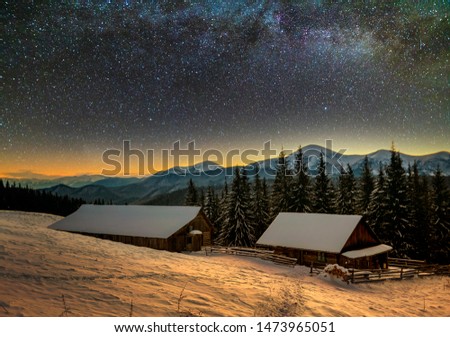Old wooden house, hut and barn, pile of firewood in deep snow on mountain valley, spruce forest, woody hills on dark starry sky and Milky Way copy space background. Mountain winter night landscape.