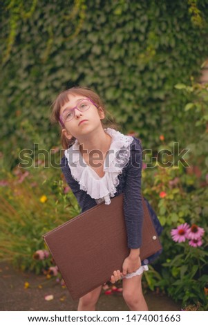 Schoolgirl with a big brown notebook on a background of greenery. Little girl 9 years old in school clothes. A girl with blond hair and a white hairpin. Blue and white clothes. Back to school