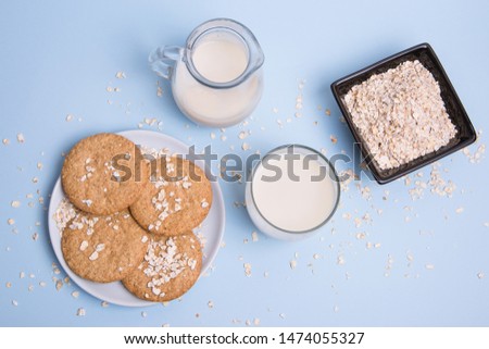 Table with a glass of vegan milk made with oat, a jar, a plate with oat biscuits and a bowl with oat flakes on a pastel blue background.