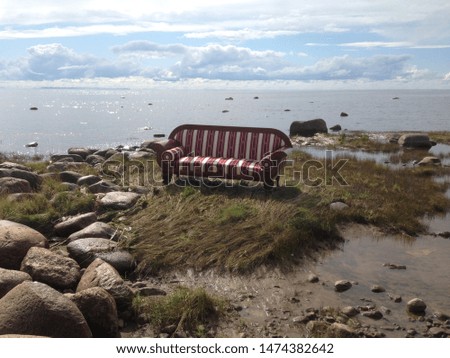 Red white lines lounge sofa or couch on the coast on green grass and rocks or stones with sea view and blue sky with clouds and quite water summer autumn season art object performance photo modern
