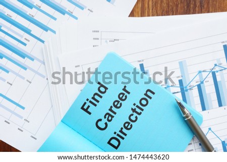 Writing note showing Find Career Direction. The text is written on a small blue notebook. Papers with graphs and colored papers, pen, wooden background are on the photo too.