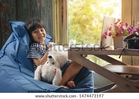 Cute Little asian Girl Sitting use Laptop at the Desk in the Living Room near window. Child Doing Homework on Computer, Browses Through Internet and Watches Cartoons. social network technology.