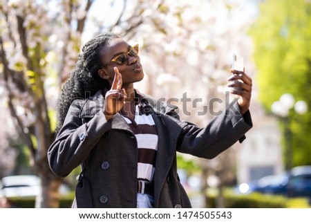 Selfie portrait of smiling young woman standing on the city street gesturing peace sign