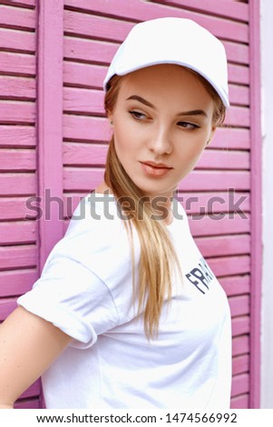 Attractive blonde dressed in a white cap, t-shirt and pants walks down the street near the pink wall on a summer day