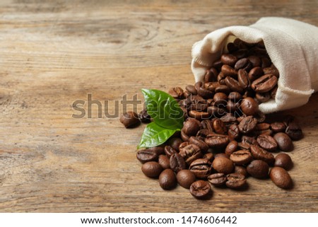 Bag of coffee beans and fresh green leaves on wooden table, space for text