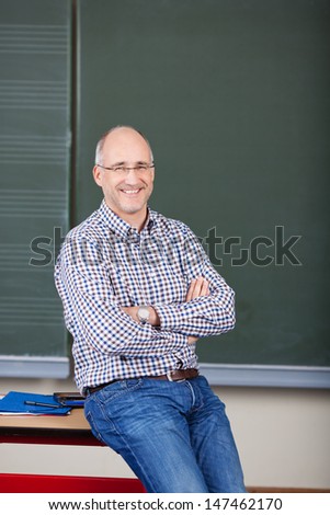 Portrait of happy male professor with arms crossed sitting on desk in classroom