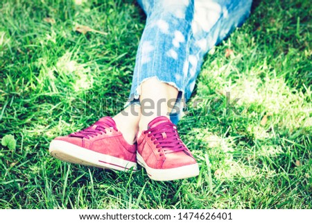 Young woman legs in sport shoes sneakers of pink suede, sitting on the grass lawn in park