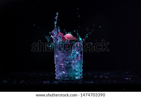 An abstract art picture presenting a pink flamingo standing in a glass full of neon colorful liquid. Black background, colors, water splash.
