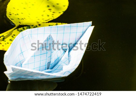 Small lonely paper boat in a cage. Notebook boat. A light sailboat floats on water. A ship in a puddle with yellow fallen autumn leaves.
