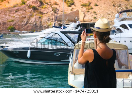 A girl in a white hat and a black T-shirt takes pictures of a bay with yachts on a telephone.