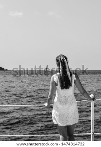 Brown haired girl wearing white dress and scarf in her hair looking out over Mediterranean turquoise water. Beautiful sunny day, clear blue sky. Black and white shot. Cannes, Côte d’Azur, France