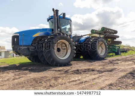 A large, multi-wheeled, blue tractor stands in a field with a green disc plow.A large, multi-wheeled, blue tractor stands in a field with a green disc plow.