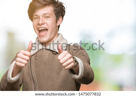 Young handsome man wearing winter coat over isolated background approving doing positive gesture with hand, thumbs up smiling and happy for success. Looking at the camera, winner gesture.