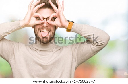 Young handsome business man wearing glasses doing ok gesture like binoculars sticking tongue out, eyes looking through fingers. Crazy expression.