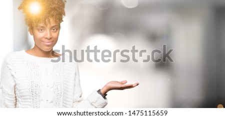 Beautiful young african american woman wearing winter sweater over isolated background smiling cheerful presenting and pointing with palm of hand looking at the camera.