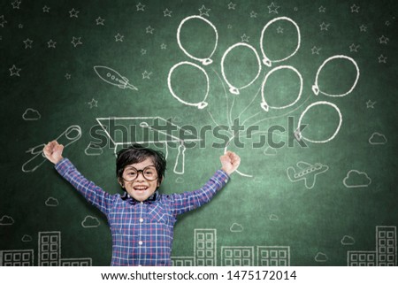 Picture of little boy looks happy while celebrating his graduation and holding a diploma and graduation cap in chalkboard background