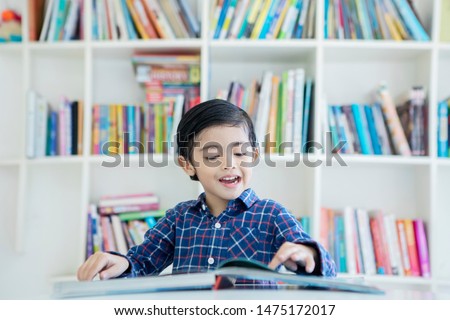 Picture of cute little boy reading a book while studying in the library with bookcase background 