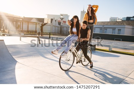 Man on bicycle with a female friend sitting on the handlebar taking selfie and another standing in back making loser hand gesture.