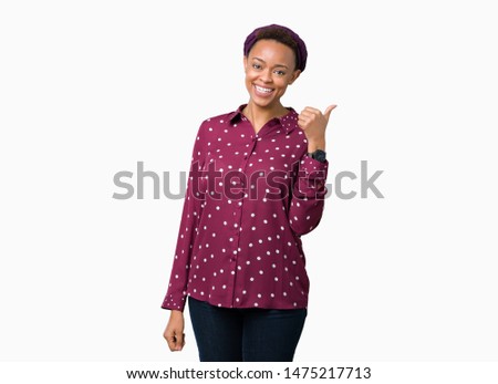Beautiful young african american woman wearing head scarf over isolated background smiling with happy face looking and pointing to the side with thumb up.