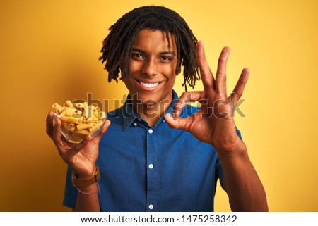 Afro american man with dreadlocks holding pasta macaroni over isolated yellow background doing ok sign with fingers, excellent symbol