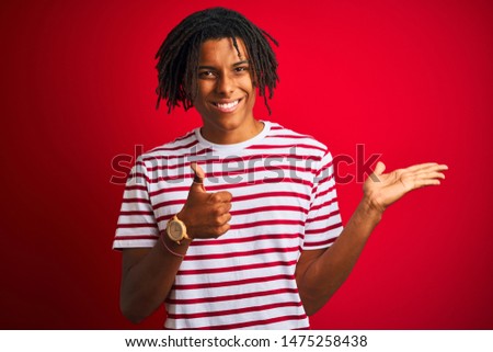 Young afro man with dreadlocks wearing striped t-shirt standing over isolated red background Showing palm hand and doing ok gesture with thumbs up, smiling happy and cheerful