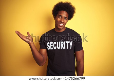 American safeguard man with afro hair wearing security uniform over isolated yellow background smiling cheerful presenting and pointing with palm of hand looking at the camera.