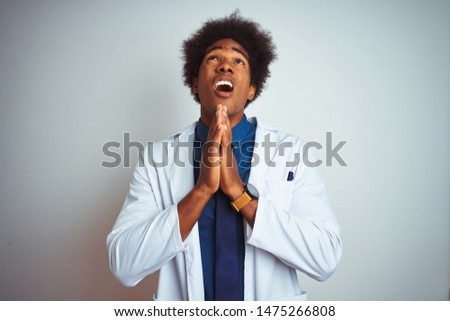 Young african american doctor man wearing coat standing over isolated white background begging and praying with hands together with hope expression on face very emotional and worried.
