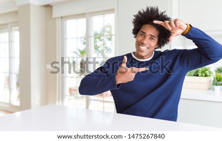 Young african american man wearing casual sweater sitting at home smiling making frame with hands and fingers with happy face. Creativity and photography concept.