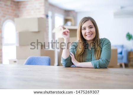 Young woman sitting on the table with cardboard boxes behind her moving to new home with a big smile on face, pointing with hand and finger to the side looking at the camera.