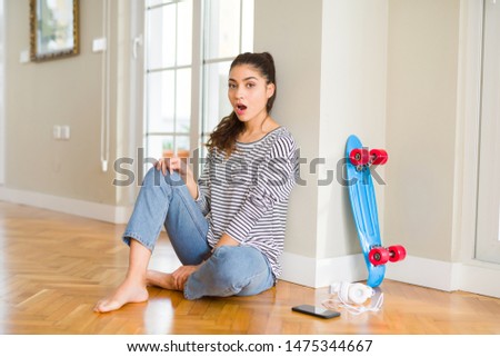 Young woman sitting on the floor using skateboard and headphones afraid and shocked with surprise expression, fear and excited face.