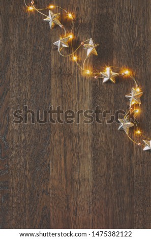 Christmas garland with golden stars lying down on wooden background lighten up by decorative lights, view from above, space for a greeting text