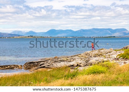 Man taking piture of Beach with rocks and vegetation and Conemara mountain in background in Carraroe, Conemara, Galway, Ireland