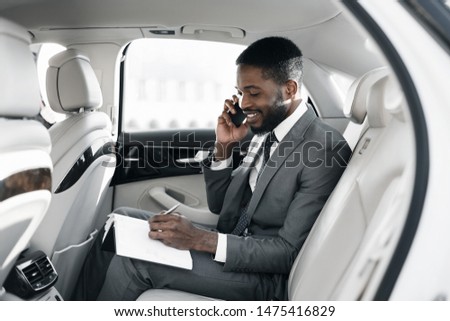 Afro businessman arranging his affairs with notepad in the car, talking on phone