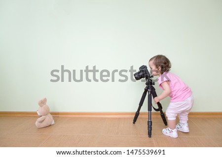 Cute funny beautiful baby photographs her bunny. The baby stands and holds a DSLR camera on a tripod against the background of a green wall. Concept novice beginner photographer, first steps.