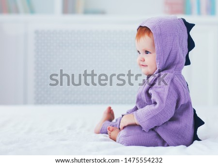 beautiful smiling, nine month old, infant baby girl in dinosaur costume, is sitting on the bed