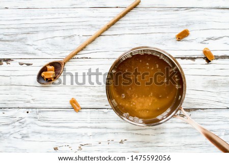 Sauce pa of melted caramel sauce with wooden spoon and candy pieces over a white wood table background. Top View.