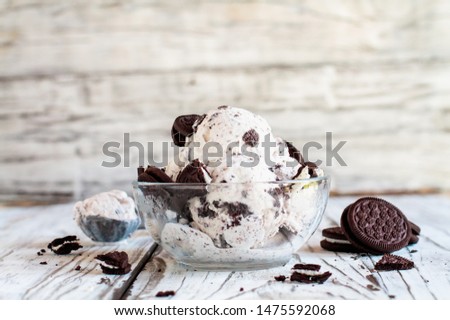 Clear glass bowl of cookies and cream ice cream. Selective focus with blurred background. Whole and crumbled cream filled cookies scattered about table. 