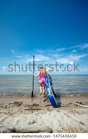 Young beautiful woman in a bright pink bikini holding a SUP board in the sea beach. Adventure girl on Standup paddleboarding with a paddle in the ocean. Back view.