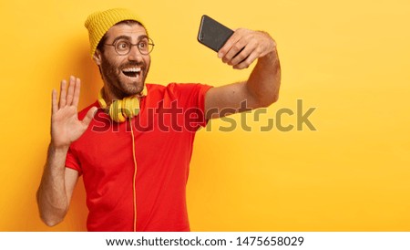 Delighted positive guy looks in mobile phone and waves palm at camera, greets someone while makes video call, smiles broadly, dressed casually, wears headphones on neck, isolated on yellow background