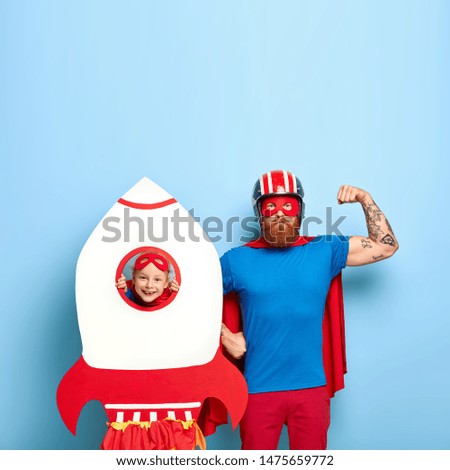 Serious dad shows biceps, dressed in headgear, blue t shirt and red cape, pretends being superhero, small child looks through paper rocket, enjoys playing with father, stand against blue wall. Family Royalty-Free Stock Photo #1475659772