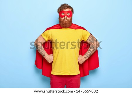 Photo of serious male in superhero costume, keeps hands on waist, possesses extraordinary talents, ready to protect our universe, isolated on blue wall. Bearded guy in red mask, cape, yellow t shirt Royalty-Free Stock Photo #1475659832