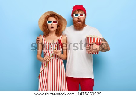 Photo of boyfriend and girlfriend have date in cinema, watch scary movie, woman in striped summer dress and straw hat holds red flower, bearded guy embraces lover, stands with basket of popcorn