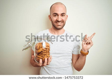 Young man holding jar of chocolate chips cookies over isolated background very happy pointing with hand and finger to the side