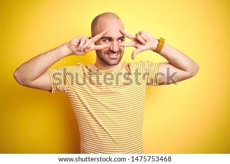 Young bald man with beard wearing casual striped t-shirt over yellow isolated background Doing peace symbol with fingers over face, smiling cheerful showing victory