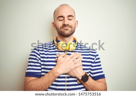 Young man listening to music wearing yellow headphones over isolated background smiling with hands on chest with closed eyes and grateful gesture on face. Health concept.