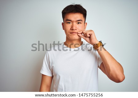 Young asian chinese man wearing t-shirt standing over isolated white background mouth and lips shut as zip with fingers. Secret and silent, taboo talking