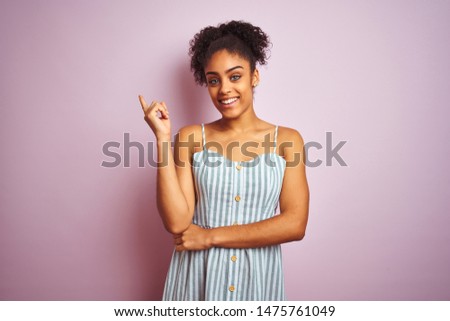African american woman wearing casual striped dress standing over isolated pink background with a big smile on face, pointing with hand and finger to the side looking at the camera.