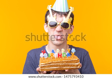 Crazy cheerful young man in glasses and paper congratulatory hats holding cakes happy birthday standing on a yellow background. Jubilee congratulations concept.