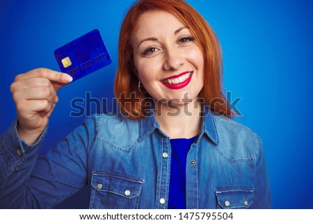 Young beautiful redhead woman holding credit card over blue isolated background with a happy face standing and smiling with a confident smile showing teeth